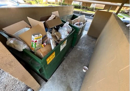 Cardboard and bags piled above green collection bins at Woodbridge Apts before service